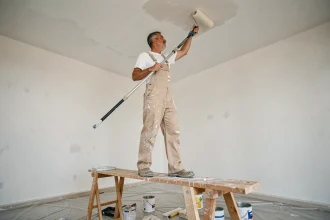 Homme en salopette peignant un plafond avec un rouleau à long manche sur un échafaudage en bois dans une pièce vide